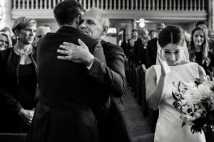 bride wipes tear as her dad hugs her soon to be husband at the end of an aisle in a church