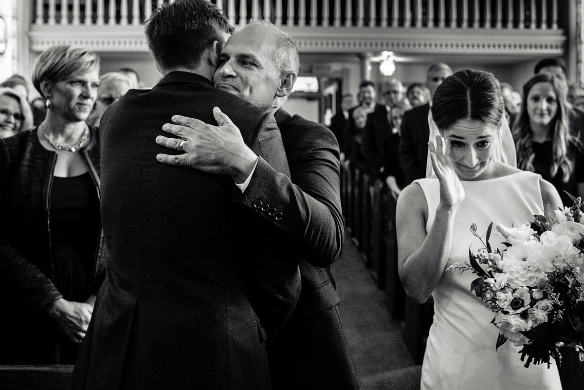 bride wipes tear as her dad hugs her soon to be husband at the end of an aisle in a church 