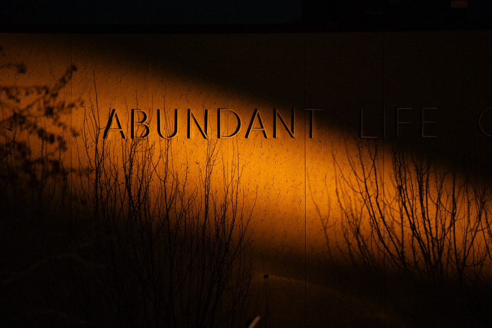 n amber security light casts a haunting glow over the words “ABUNDANT LIFE” on a wall, partially obscured by the silhouettes of bare winter branches — taken at the scene of a December school shooting.