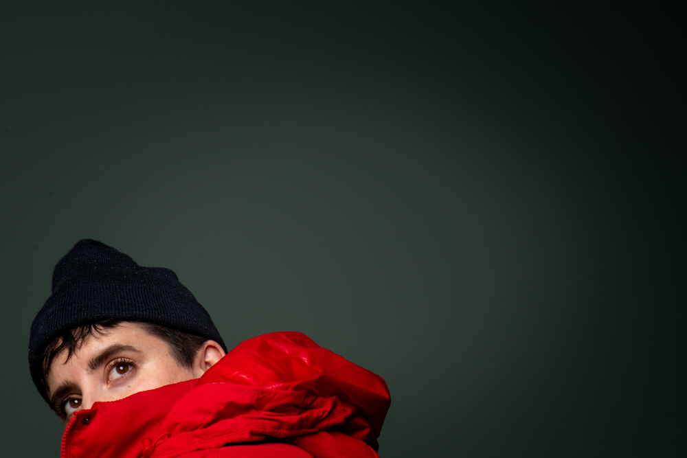 Portrait of a person in a red puffer jacket and dark beanie, glancing sideways with only their eyes and part of their face visible against a dark green backdrop.