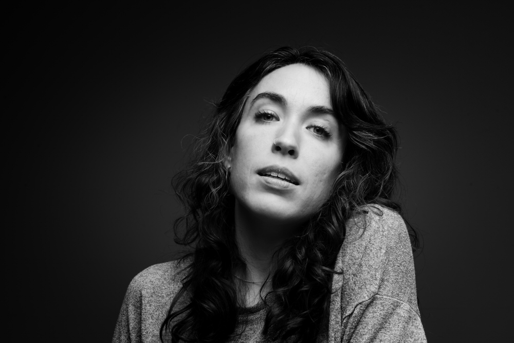 Black and white studio portrait of a woman with long wavy hair and a nose ring, wearing a loose sweater and gazing softly at the camera with a slight tilt of her head.