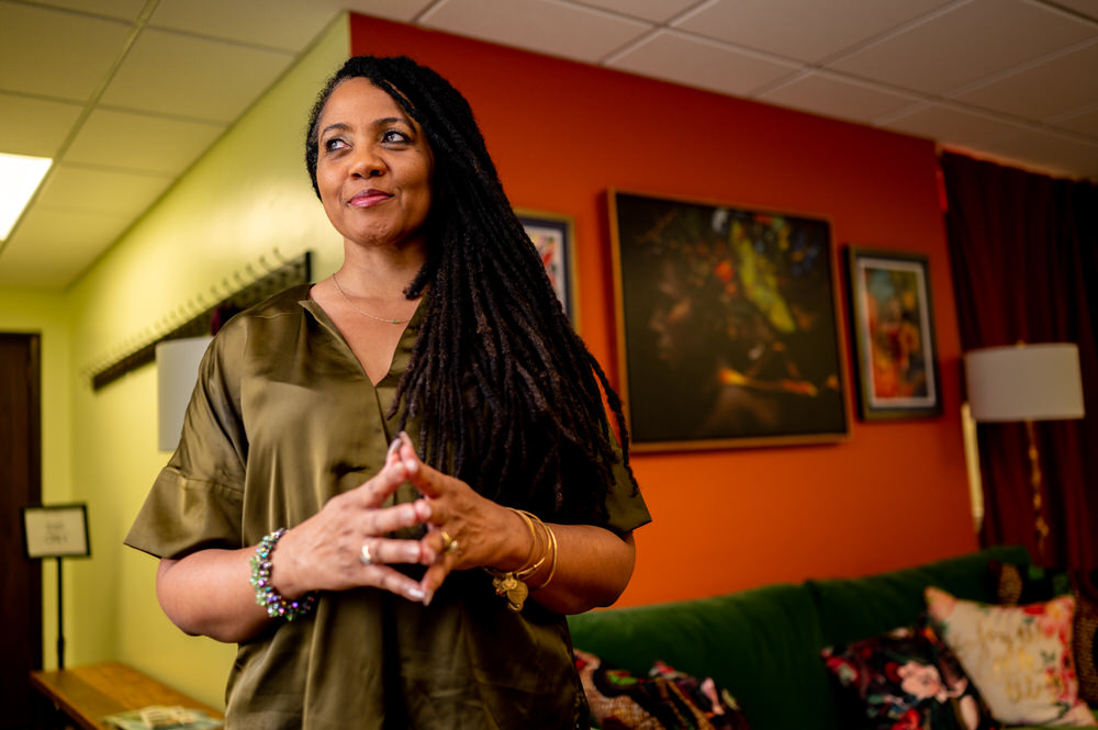 A confident woman with long locs stands in a brightly colored room, wearing a satin olive blouse, hands loosely clasped, with colorful art and pillows behind her.