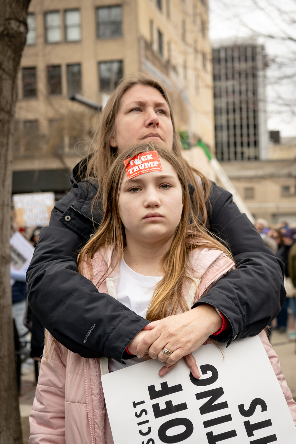 A woman embraces a young girl during a protest. The girl has a sticker on her forehead that reads “F*** TRUMP” and holds a partially visible protest sign. They stand in front of city buildings with other demonstrators blurred in the background.