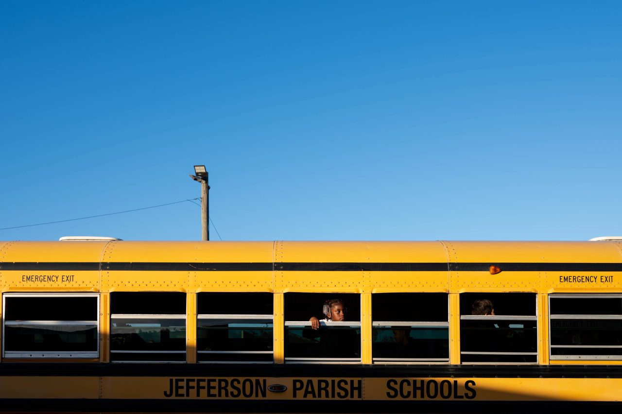 A yellow Jefferson Parish school bus is pictured against a clear blue sky. A child with headphones looks out of an open window, resting one arm on the sill, while other students are partially visible inside the bus.