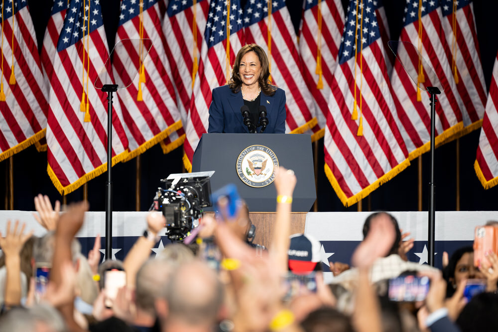 Vice President Kamala Harris smiles at the podium during her campaign kickoff rally in West Allis, Wisconsin, with American flags behind her and a cheering crowd in the foreground.