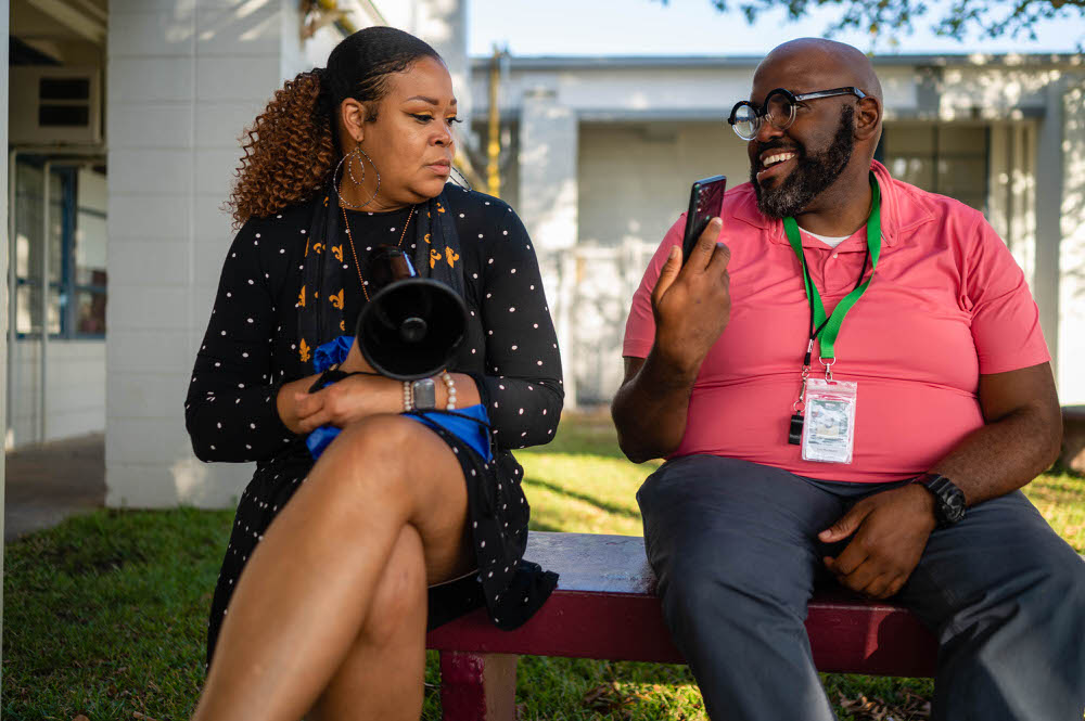A math interventionist in New Orleans shares a moment with a colleague, smiling as he shows something on his phone.