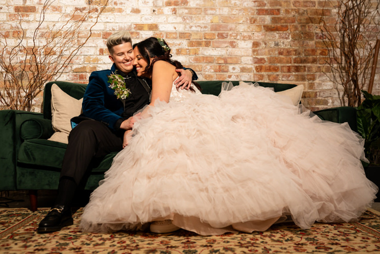 Nichole+Monica_0207 Two newlywed brides share a joyful embrace on a velvet green couch at Garver Feed Mill, framed by warm brick walls and soft natural light.